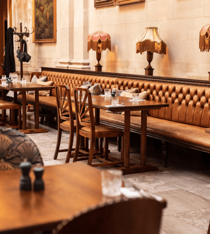 A cozy restaurant interior with brown leather tufted seating, wooden tables and chairs, vintage lamps on the wall, and framed paintings, creating a warm, inviting atmosphere.