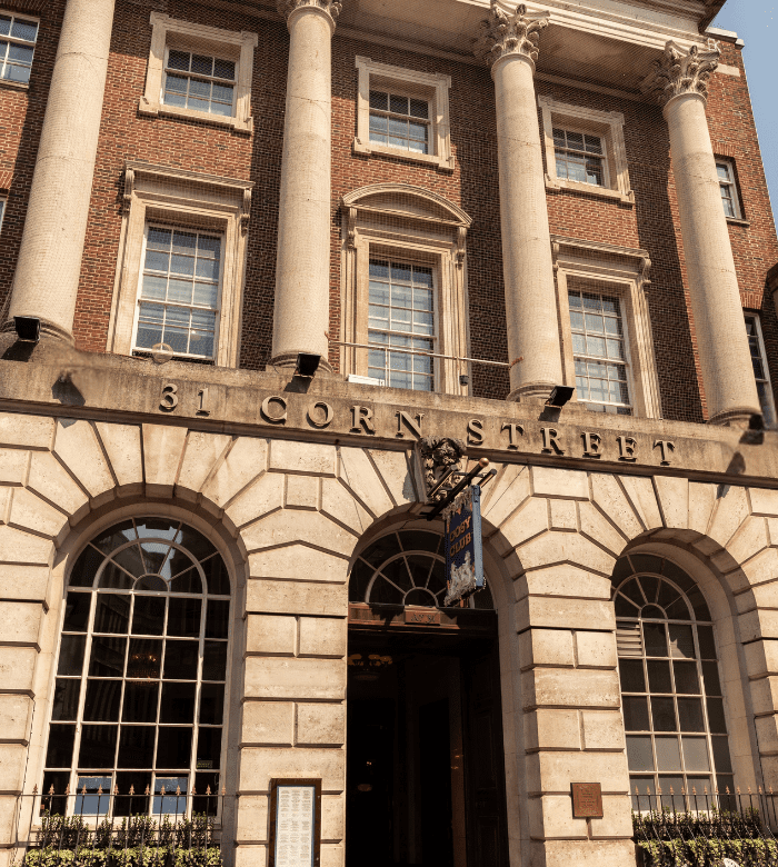 The entrance of a historic brick building with large columns, arched windows, and "31 Corn Street" engraved above the doorway. A blue and gold sign hangs near the entrance.