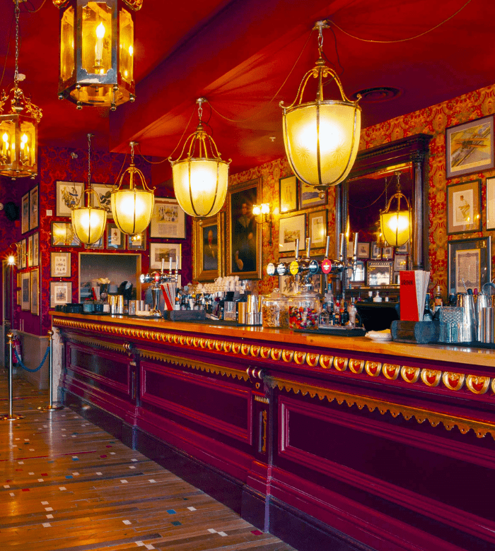 A vintage-style bar with ornate red decor, gold accents, large hanging lanterns, framed pictures on the wall, and various bottles and taps lined up on the counter. The atmosphere is warm and elegant.