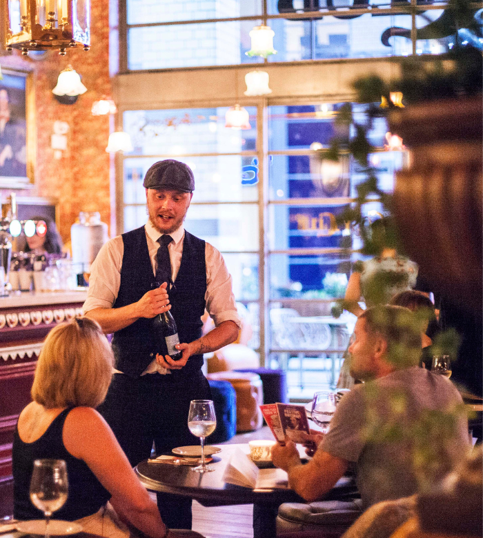 A server in a vest and flat cap opens a wine bottle at a cozy, warmly lit restaurant as two seated guests watch. The bar and hanging lights create a welcoming atmosphere in the background.