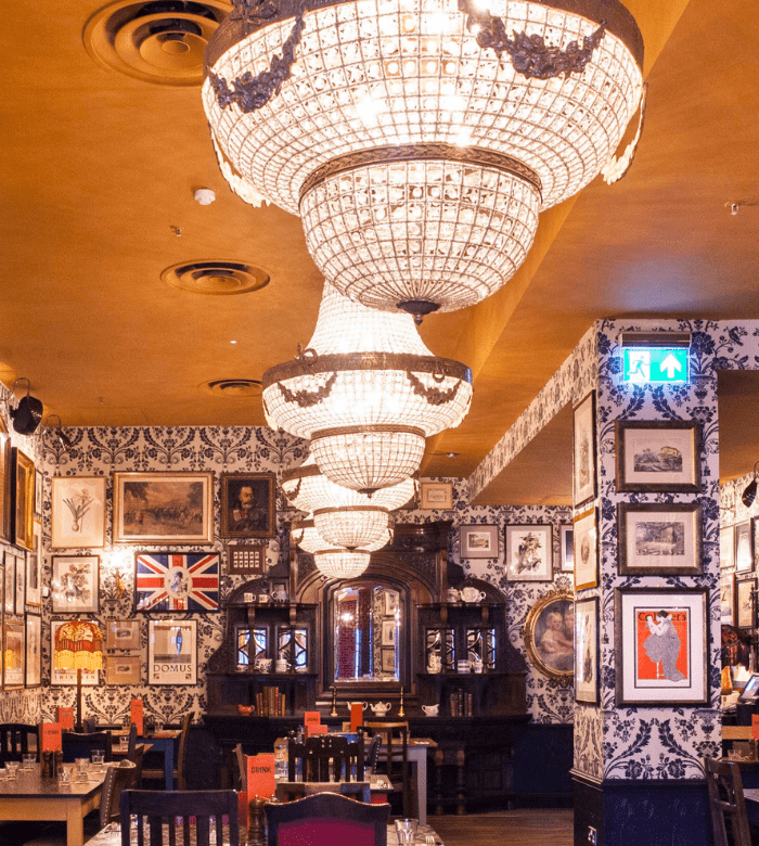 A warmly lit restaurant with ornate crystal chandeliers, patterned wallpaper, framed art and photographs, a prominent Union Jack flag, and neatly set tables with wine glasses.