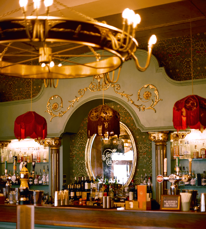 A vintage-style bar with ornate gold decor, a large round mirror, shelves stocked with bottles, and red fringed lampshades hanging over the counter. A chandelier with glowing lights is visible above.