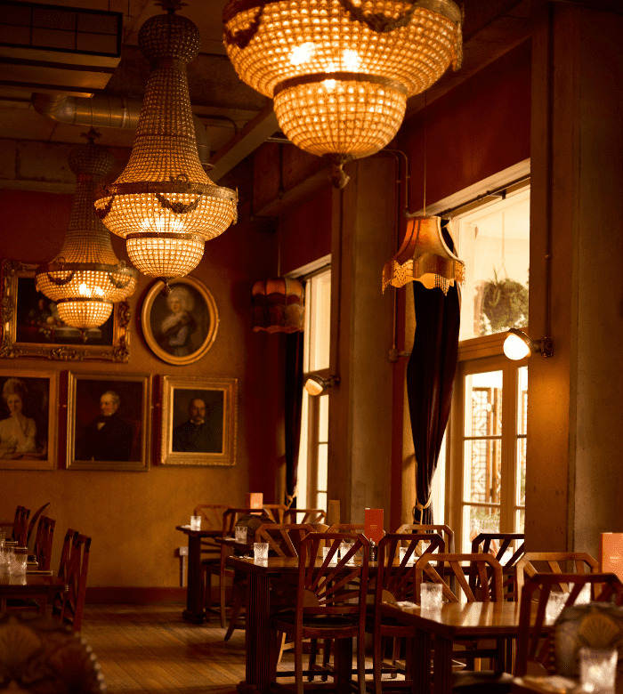 A warmly lit vintage restaurant with ornate chandeliers, portraits on the walls, wooden tables and chairs, and large windows with draped curtains letting in natural light.