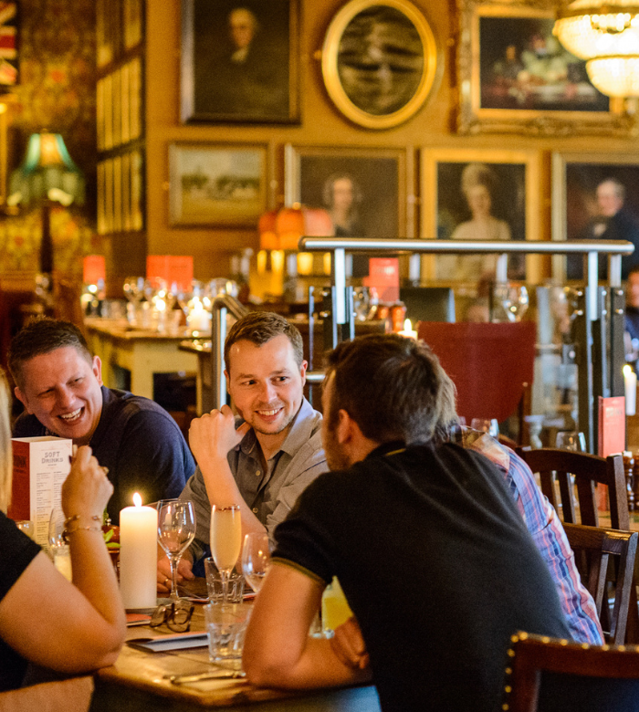 A group of people sit around a candlelit table in a warmly lit restaurant, smiling and talking. The background features framed paintings and decorative lighting, creating a cozy, inviting atmosphere.