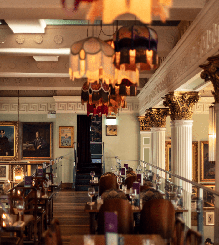 Elegant restaurant interior with ornate columns, vintage-style hanging lamps, framed paintings on the walls, and tables set with glasses and menus, creating a warm and sophisticated atmosphere.