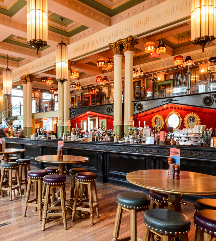 A spacious, elegant bar with high ceilings, ornate columns, hanging lamps, wooden floors, round tables, and upholstered stools. The bar back features mirrors, bottles, and red accents, with a mezzanine level above.