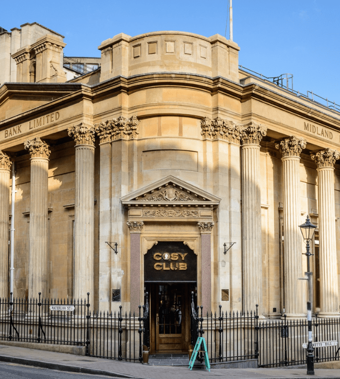 A grand neoclassical building with tall columns and ornate details houses the "Cosy Club" restaurant; "Midland Bank Limited" is inscribed above, and a black fence runs along the front.