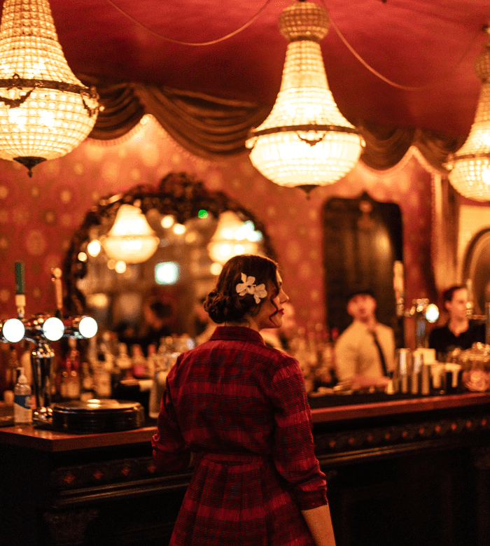 A woman in a red plaid dress with a white flower in her hair stands at an ornate bar, lit by large chandeliers. The bar has a vintage, elegant decor with gold accents and a large mirror behind it.