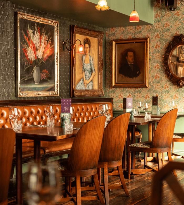 A cozy restaurant with wooden tables and chairs, brown tufted leather bench seating, floral wallpaper, framed portraits, and soft ambient lighting. Dining tables are set with glassware and menus.