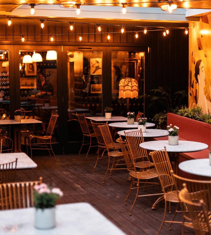 Outdoor café patio with marble-topped tables, gold chairs, string lights overhead, and potted flowers on each table. Walls have warm lighting and painted murals, creating a cozy, inviting atmosphere.
