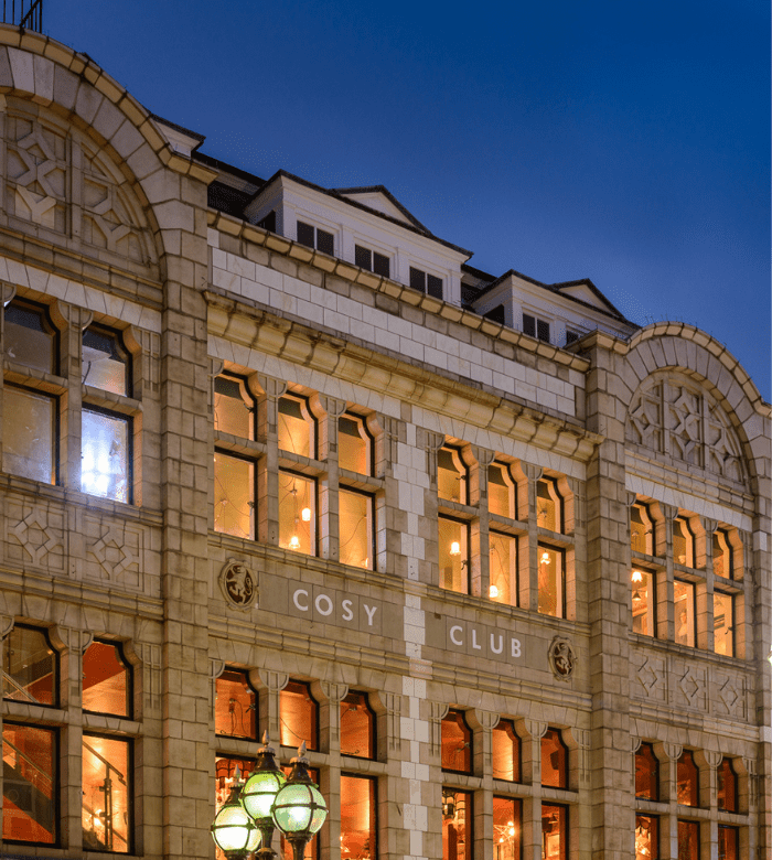 A historic stone building with large arched windows lit from within at dusk. The sign “COSY CLUB” is displayed above the first floor, and ornate street lamps are visible in front.