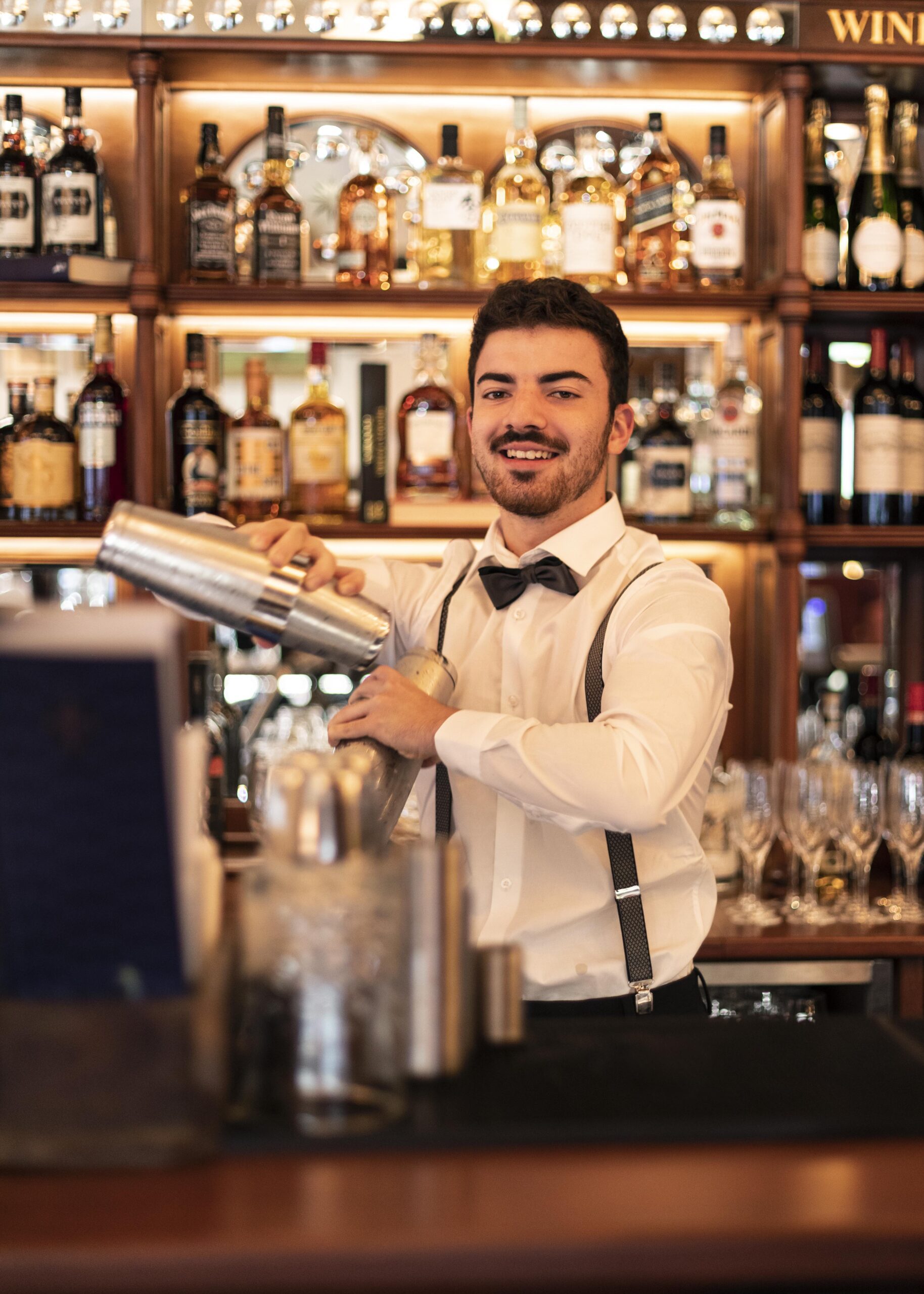 A bartender in a white shirt, bow tie, and suspenders smiles while shaking a cocktail shaker behind a bar stocked with bottles and glassware.