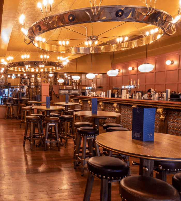 A stylish bar with round wooden tables and barstools, ornate lighting fixtures, a polished wooden floor, and a long bar with decorative panels and various drinks on display.