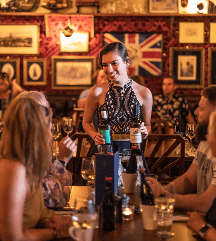 A smiling waitress in a patterned dress serves drinks to a group of people seated at a lively, warmly-lit restaurant with vintage decor and a large Union Jack flag on the wall.