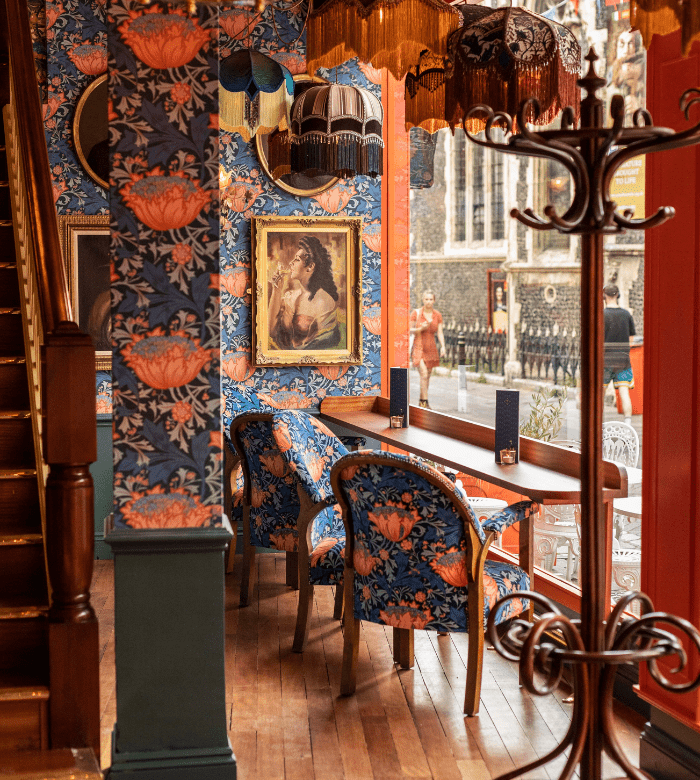 An ornately decorated café interior with floral wallpaper, matching upholstered chairs, framed art, and vintage lamps. Wooden floors and a stairway are visible, with large windows overlooking a street scene outside.