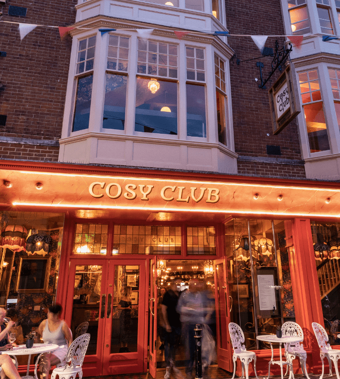 The entrance of Cosy Club, a warmly lit restaurant with outdoor seating, ornate metal chairs and tables, and large bay windows above. People are seated and entering the vibrant, inviting venue.