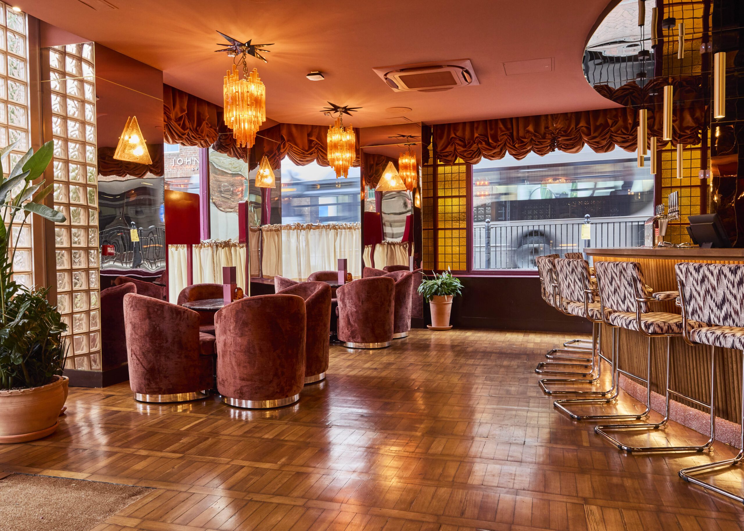 A retro-style lounge with plush velvet chairs, patterned barstools, wood flooring, gold accents, crystal chandeliers, large windows with curtains, and potted plants, creating a warm, inviting atmosphere.