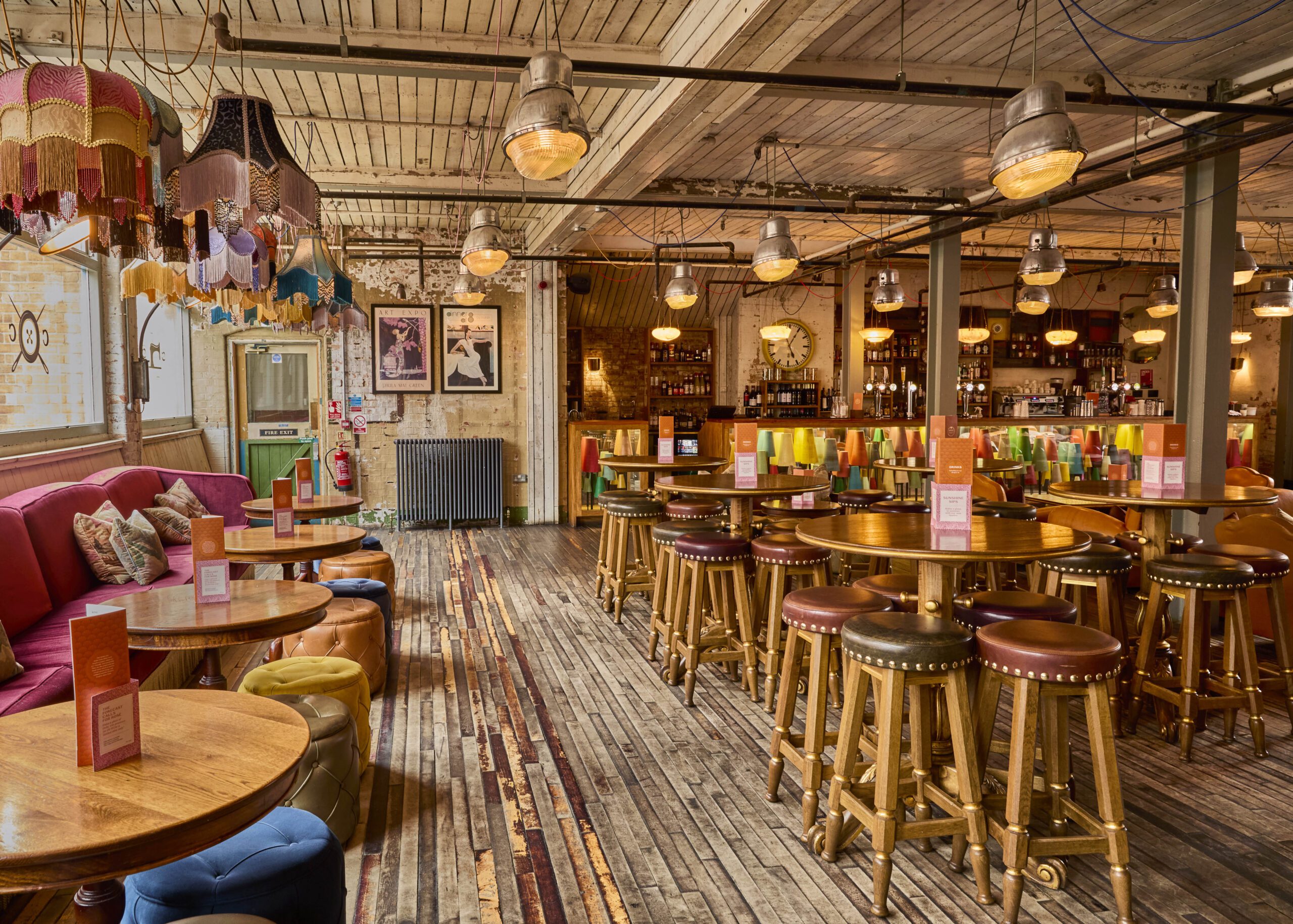 A cozy, eclectic bar with wooden floors and ceilings, round tables with stools, colorful lampshades, and a vibrant bar area in the background. Menus are placed on each table. The atmosphere is warm and inviting.