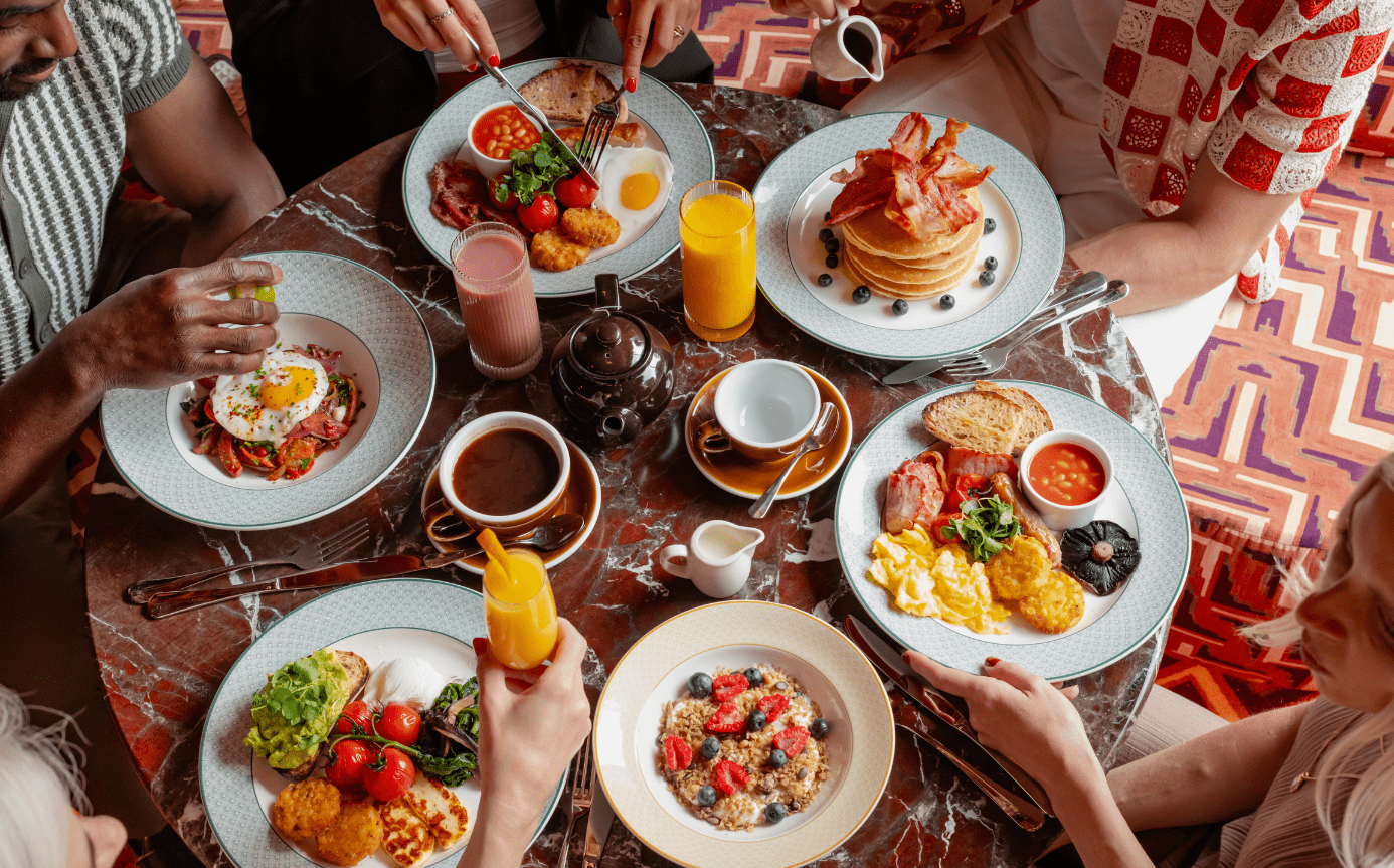Four people sit around a table with plates of breakfast foods—eggs, pancakes, bacon, toast, fruit, and drinks like orange juice and coffee—sharing a meal together.