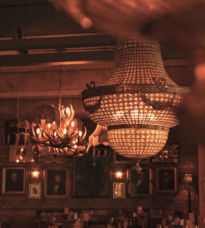 A close-up of an ornate crystal chandelier hanging in a dimly lit room with vintage lamps, framed portraits, and antler decor in the background.