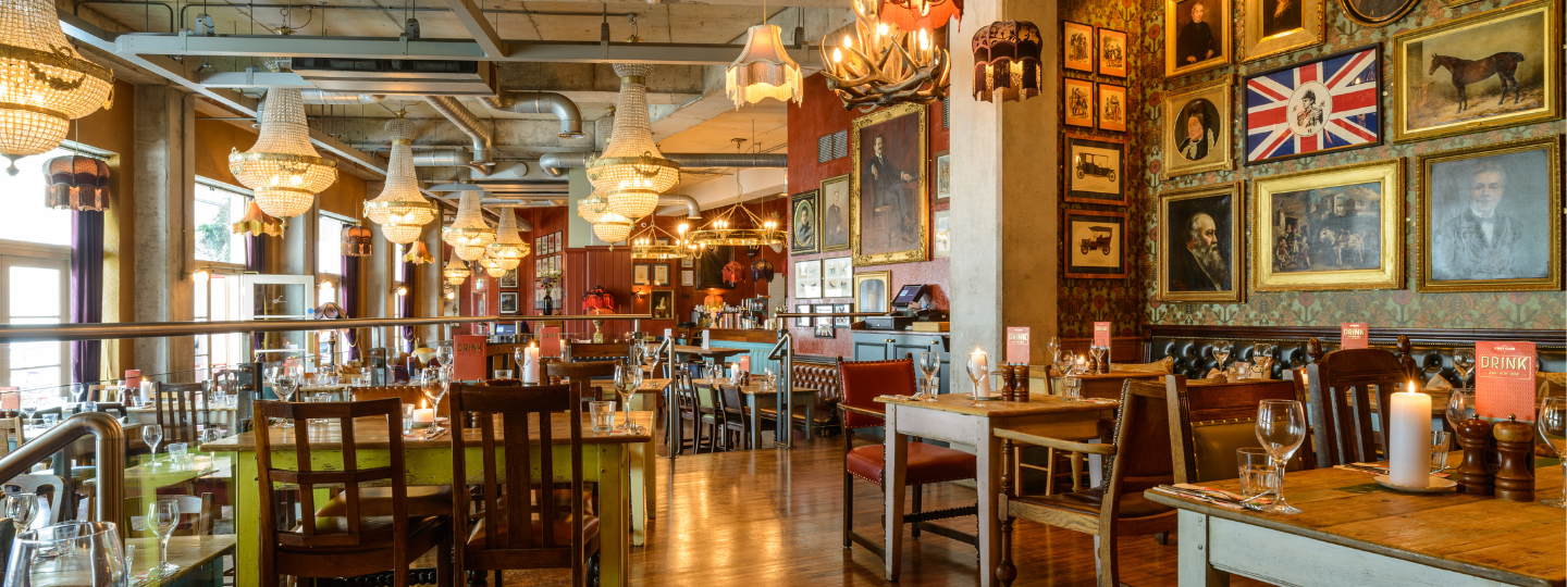 Eclectic restaurant interior with wooden tables and chairs, chandeliers, and framed portraits on patterned walls. Soft lighting, candles, and place settings create a cozy, vintage atmosphere.