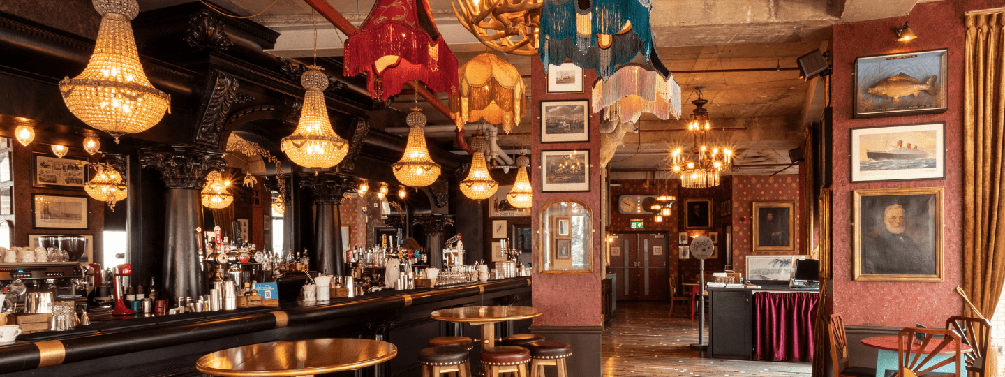 An ornate pub interior with vintage chandeliers, eclectic hanging lampshades, framed artwork on red walls, dark wood bar, and round tables with stools, creating a cozy and nostalgic atmosphere.