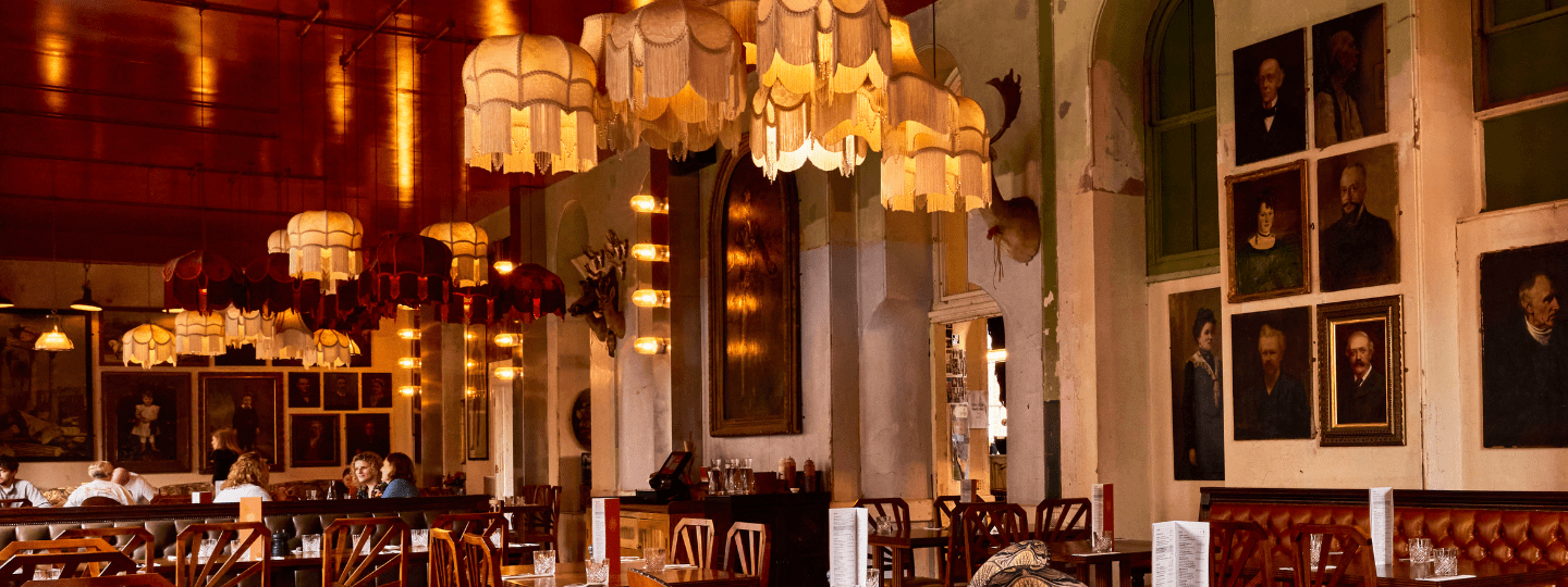 Dimly lit vintage restaurant with ornate hanging lamps, framed portraits on the walls, wooden tables and chairs, and a few people seated, creating a cozy, nostalgic atmosphere.