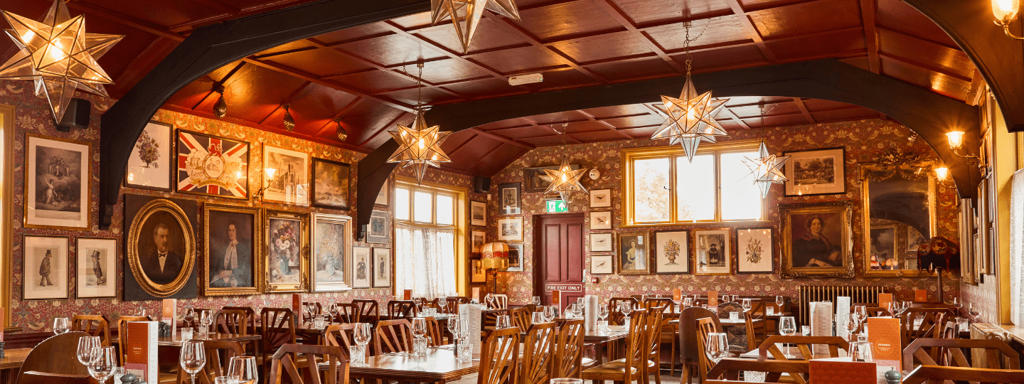 A warm, vintage-style restaurant dining room with wooden tables and chairs, star-shaped pendant lights, and walls covered in framed artwork and portraits, illuminated by natural light from large windows.