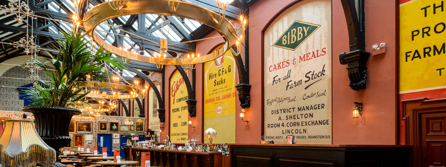 A stylish restaurant with large wall advertisements, vintage decor, circular chandeliers, wooden tables, and a high glass ceiling, creating a retro yet elegant atmosphere.
