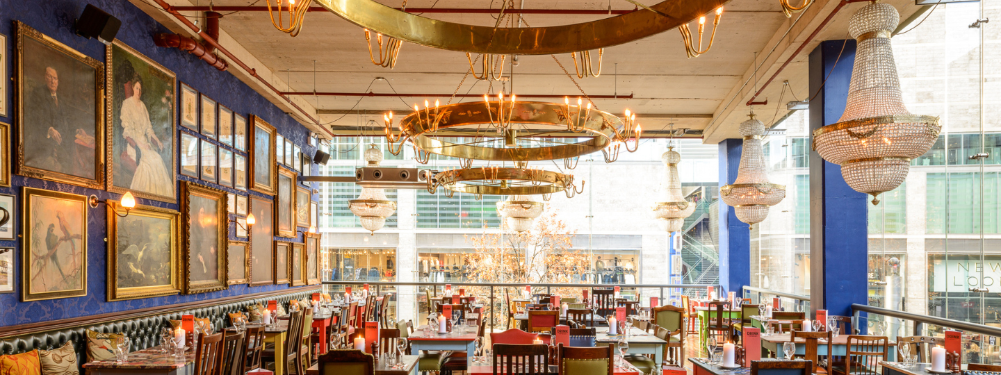 A spacious, elegant restaurant with eclectic decor, large chandeliers, gold-framed paintings on blue walls, and large windows overlooking a modern shopping area. Tables and chairs are arranged neatly throughout the space.