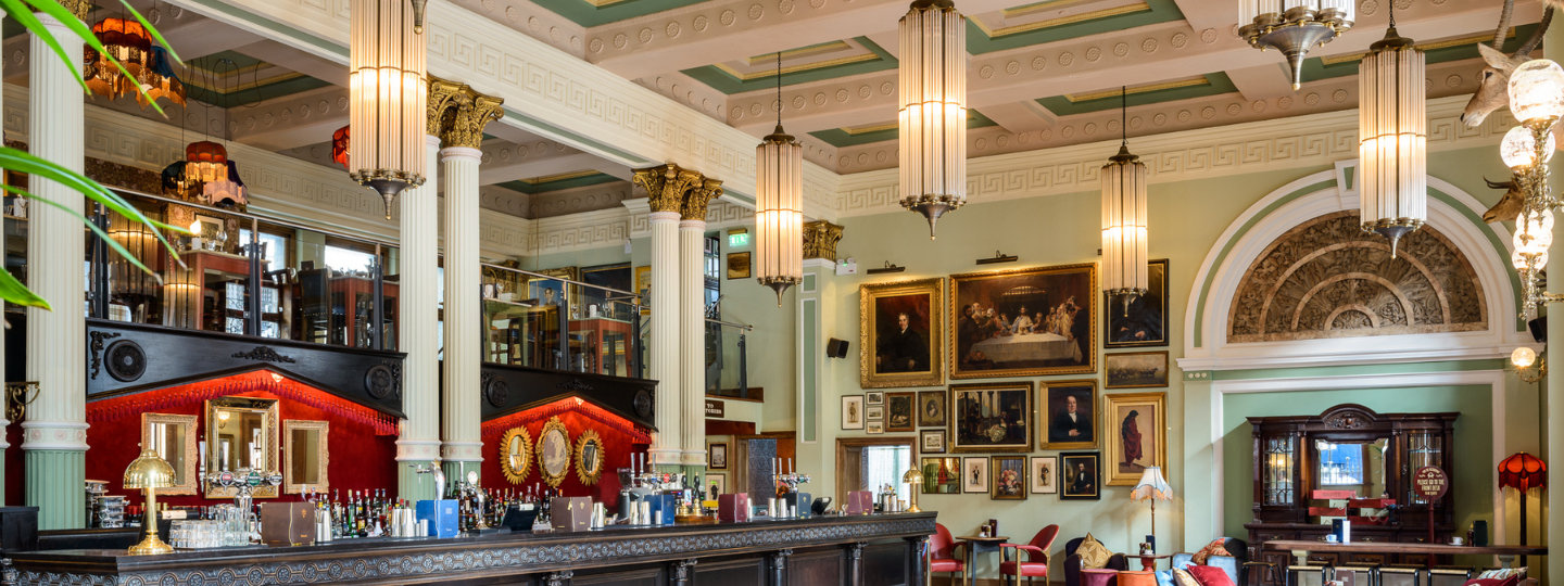 Elegant bar with tall columns, ornate ceilings, and large hanging lights. Paintings and portraits decorate the pastel-green walls. The long bar has bottles and glasses, with red accents and plush seating throughout the room.
