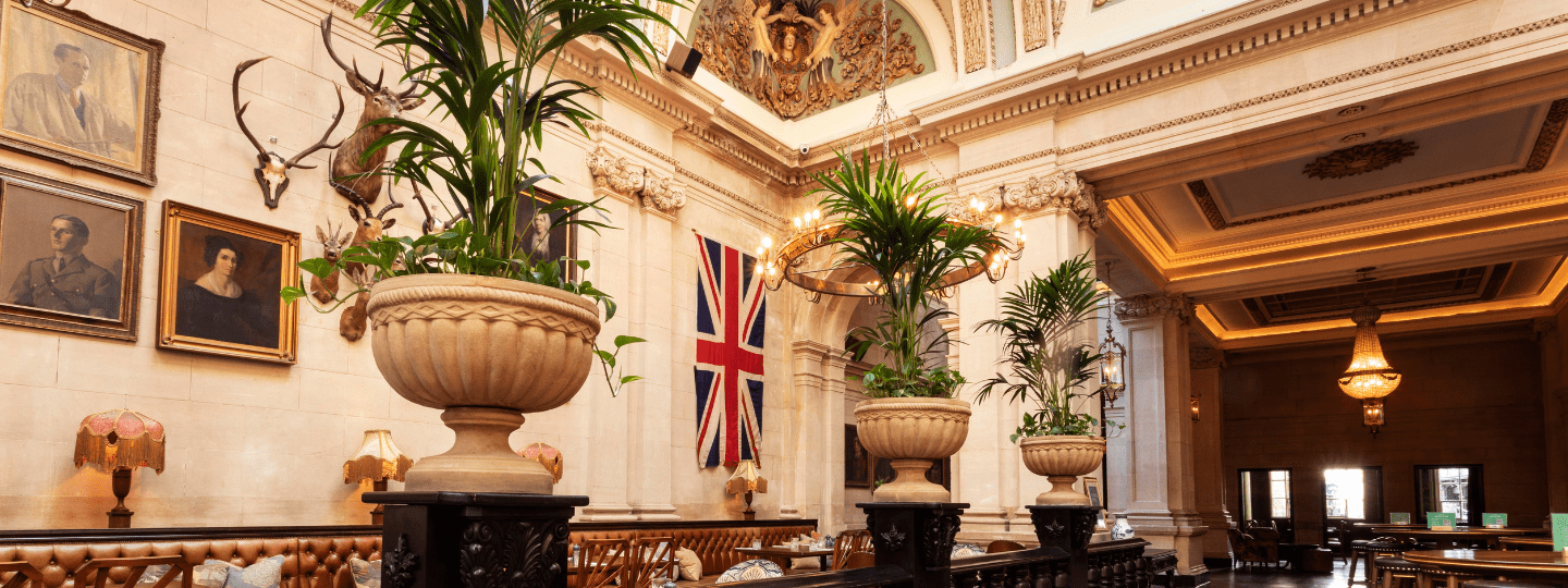 A grand, elegant interior with tall potted plants, chandeliers, vintage lamps, portraits, a large British flag, and high ornate ceilings in a lavish lounge or dining area.