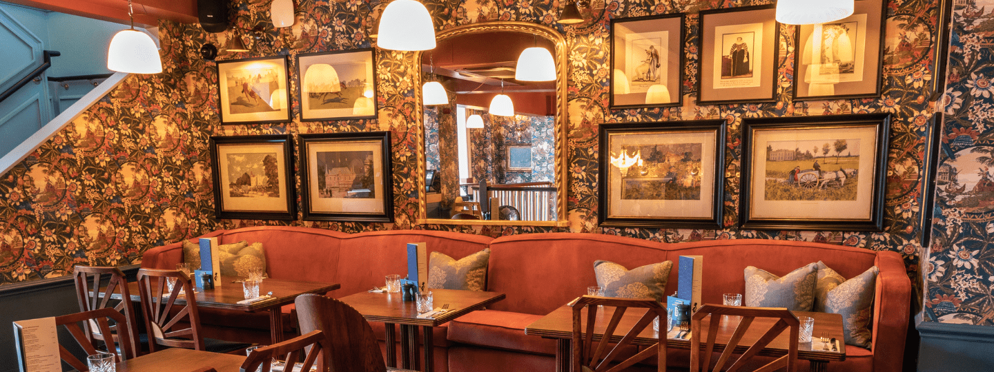 A cozy restaurant interior featuring a long red sofa with patterned cushions, wooden tables and chairs, floral wallpaper, framed art on the walls, and hanging pendant lights creating a warm atmosphere.