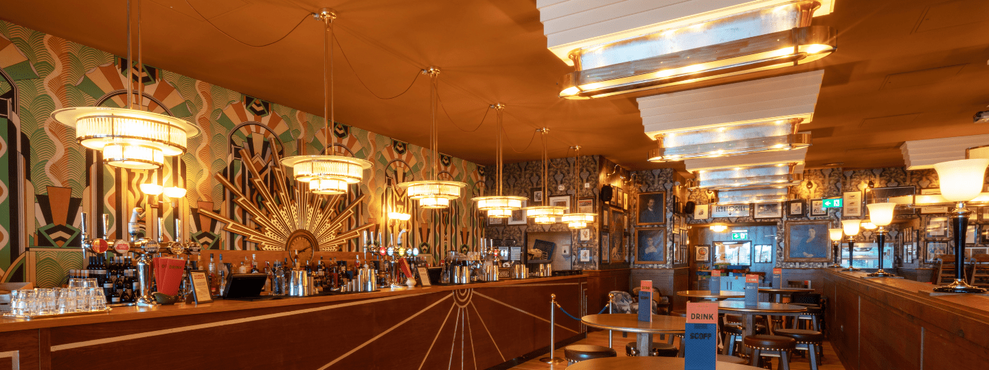 Art Deco-style bar interior with patterned wallpaper, large decorative light fixtures, a long wooden bar counter with various bottles, small round tables, and framed pictures on the walls. The space is warmly lit.