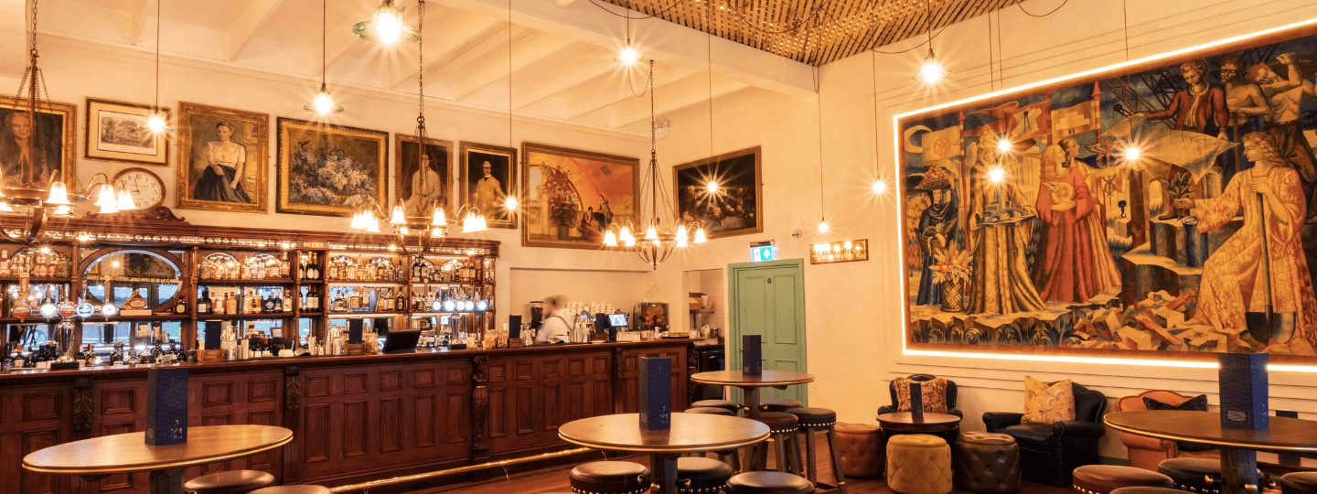 Elegant bar with wooden counters, round tables, and stools, illuminated by hanging lights. Large paintings and portraits decorate the walls, creating a warm and artistic atmosphere.