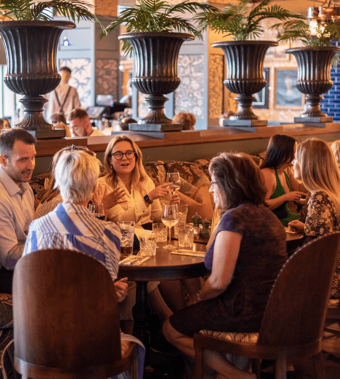 A group of people sit around a round table at a stylish restaurant, talking and enjoying drinks. Large decorative vases with plants are in the background, and the setting appears lively and social.