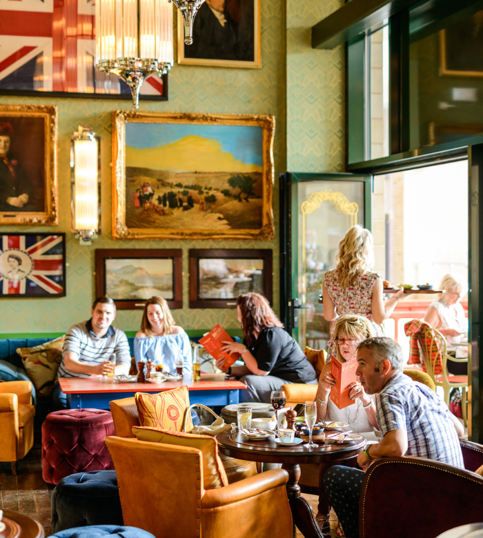 People relax and chat in a cozy, colorful café with British-themed decor, including Union Jack flags and landscape paintings. Some enjoy drinks and food at tables, while a woman stands near the open door.