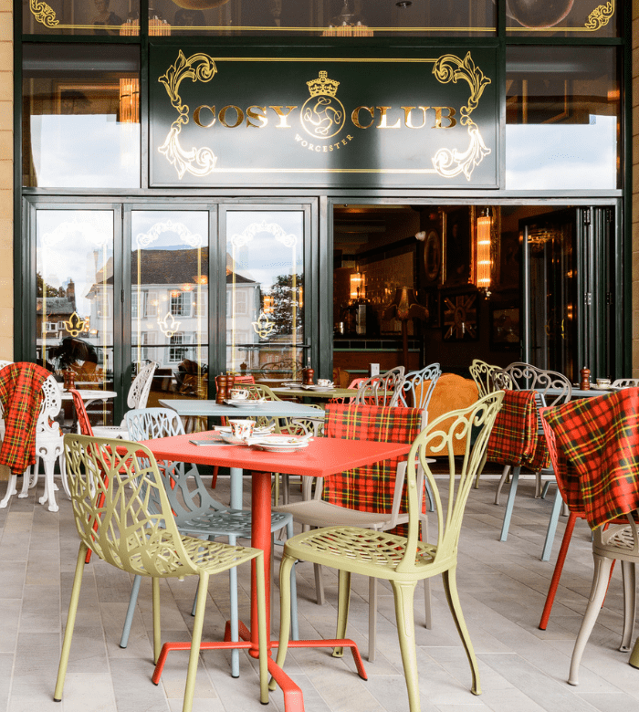Outdoor seating area of a café called Cosy Club, featuring decorative metal chairs, tables, and red tartan blankets. The entrance has large glass doors and a stylish sign above them.