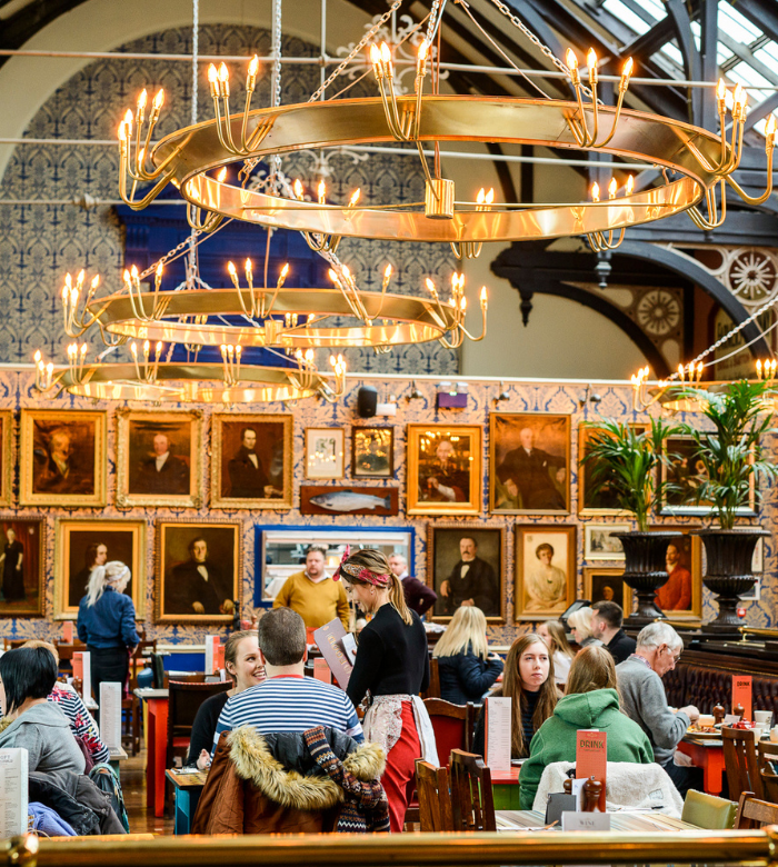 A busy café filled with people dining beneath large, circular chandeliers. The walls are decorated with framed portraits and patterned tiles, and natural light streams in from tall windows above.