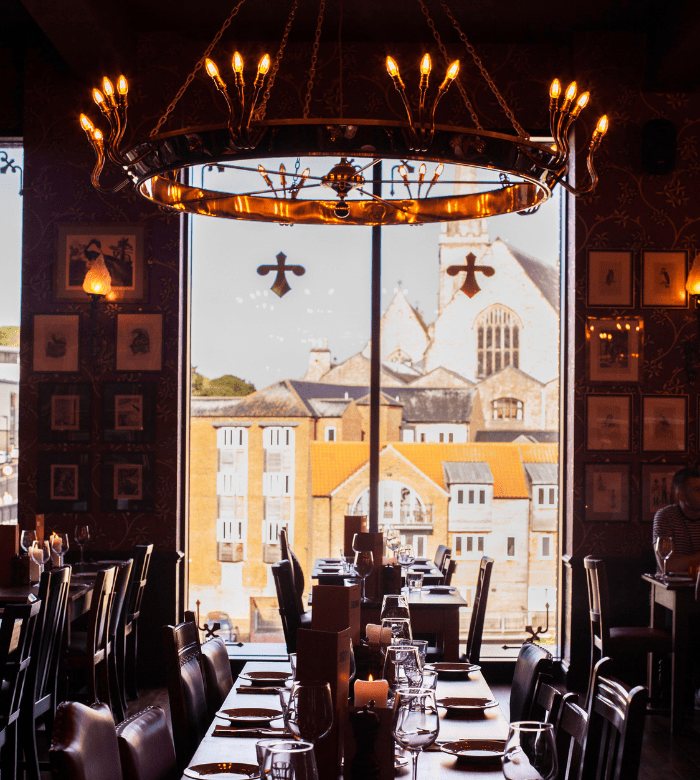 Elegant restaurant interior with a large circular chandelier, set tables, and a window offering a view of historic buildings and a church outside. Warm lighting creates a cozy atmosphere.