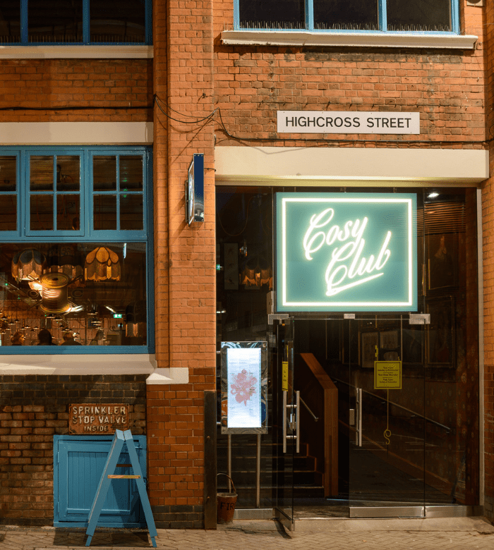 A neon "Cosy Club" sign glows above the entrance to a brick building on Highcross Street. Blue window frames, a blue chair, and warmly lit interior details are visible.