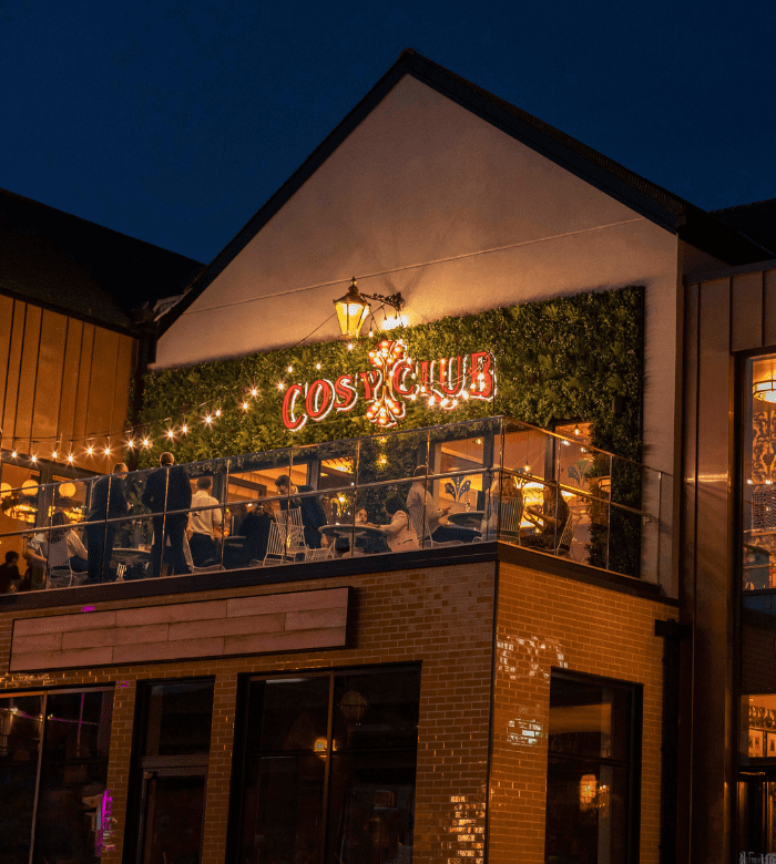 A warmly lit restaurant called Cosy Club with outdoor seating on a balcony, string lights, and people dining at night. The building has a green wall with the neon sign illuminated.