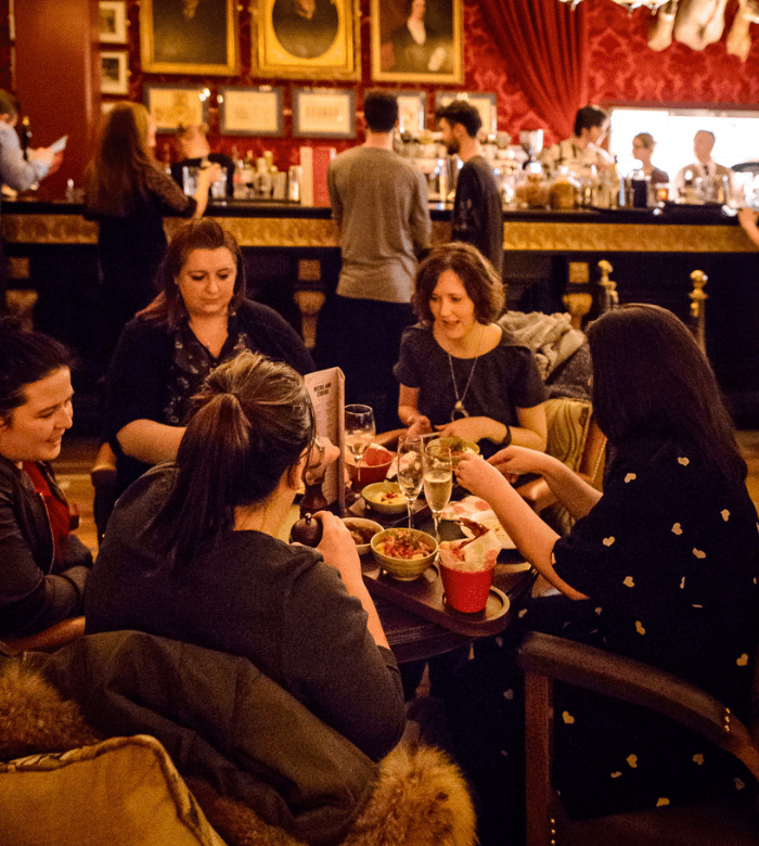 Five women are seated around a small table in a warmly lit, elegant bar, sharing drinks and food. The background shows a busy bar with people, ornate decor, and framed portraits on red-patterned walls.
