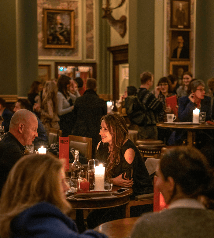 A woman and a man sit at a candlelit table in a busy, warmly lit restaurant, surrounded by other diners and decorated with framed portraits and elegant furnishings.