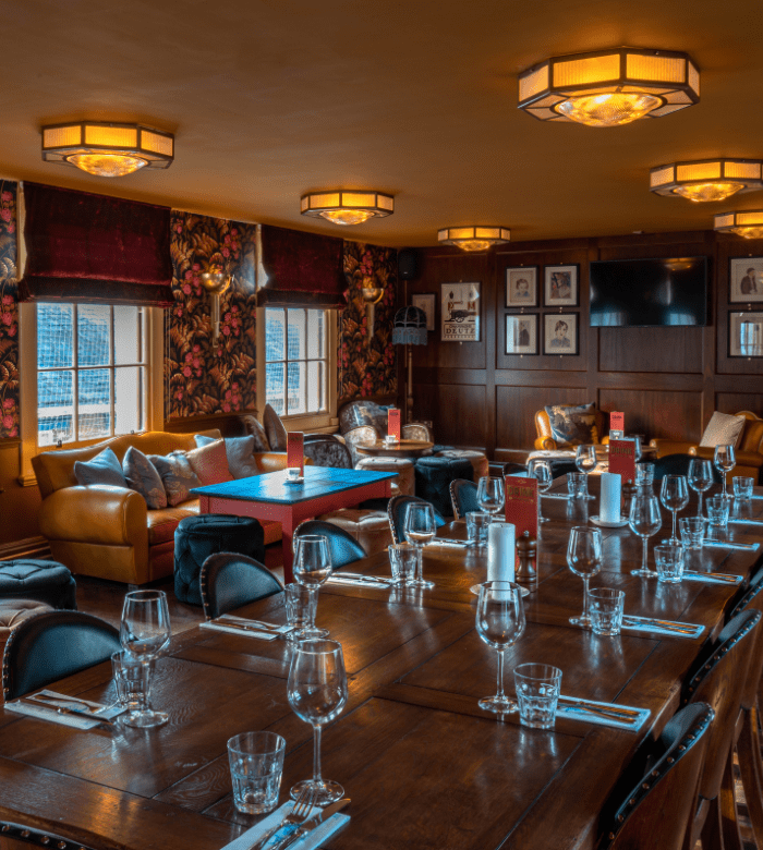 A cosy dining room with a long wooden table set with glasses, cutlery, and candles. Leather sofas, patterned wallpaper, and framed art create a warm, inviting atmosphere under soft ceiling lights.