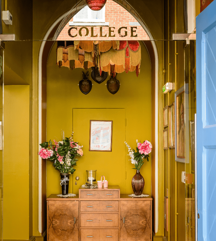 A hallway with gold walls and an arched ceiling displays the word "COLLEGE" above. A wooden sideboard holds flower arrangements and a glass water dispenser, with framed pictures and decorative tassels on the wall above.