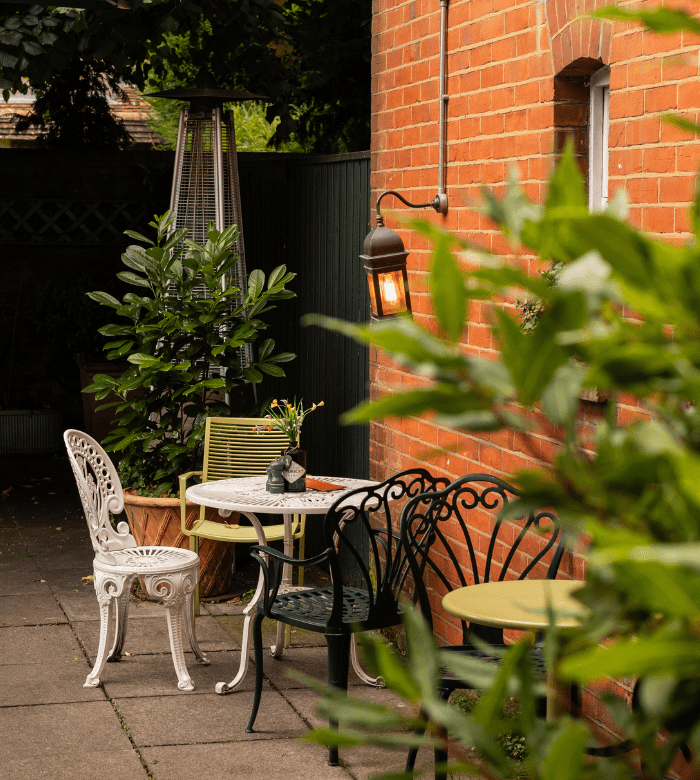 A small patio with a round table, assorted metal chairs, potted plants, and a lit wall lantern beside a red brick building. Green foliage frames the foreground.