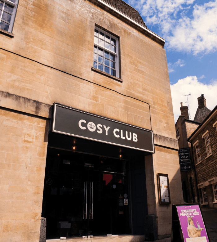 A tan brick building with a black sign reading "COSY CLUB" above the entrance. There's a pink sandwich board outside, and the sky is partly cloudy. The street and neighboring buildings are visible.