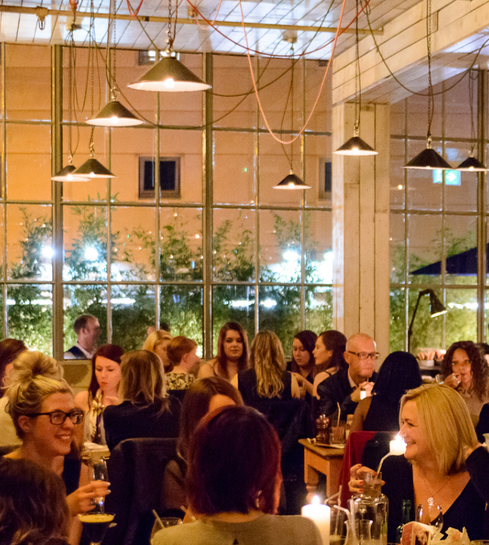 A cozy, warmly lit restaurant filled with people dining and conversing at tables. Large windows reveal a softly illuminated outdoor area. Hanging lights and candles add to the inviting atmosphere.