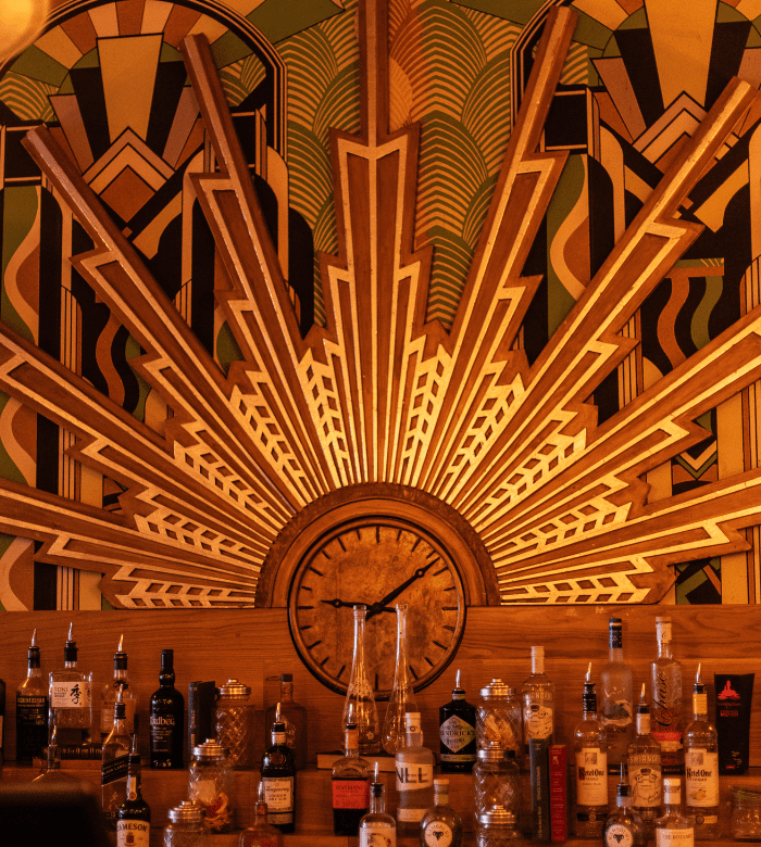 Art Deco-style bar with a vibrant sunburst mural and geometric patterns behind a wooden bar stocked with assorted liquor bottles and a central clock. Warm, golden lighting creates a vintage, inviting atmosphere.